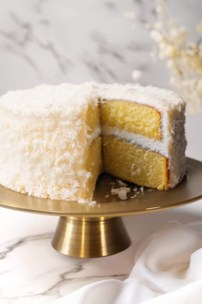 A Coconut Lovers Dream Cake with Coconut Buttercream Frosting sits elegantly on a gold cake stand. A slice is cut out, revealing the moist yellow interior. The background features a marble surface and soft, out-of-focus dried flowers, making it a perfect centerpiece for any occasion.