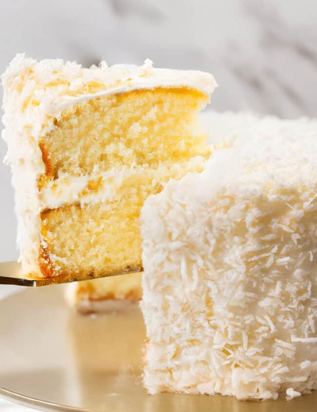 A close-up of a Coconut Lovers Dream Cake with a slice being lifted out. The cake has a light yellow interior with layers of coconut buttercream frosting, and the outer surface is covered in shredded coconut flakes. The cake is displayed on a gold cake stand with a marble background.