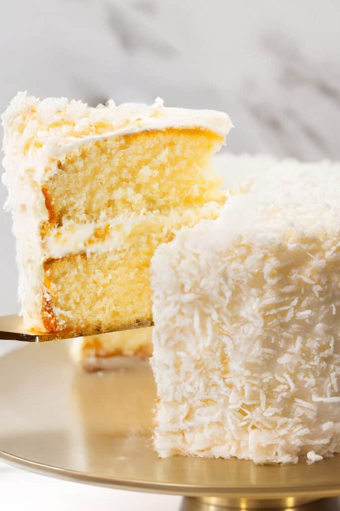 A close-up of a Coconut Lovers Dream Cake with a slice being lifted out. The cake has a light yellow interior with layers of coconut buttercream frosting, and the outer surface is covered in shredded coconut flakes. The cake is displayed on a gold cake stand with a marble background.