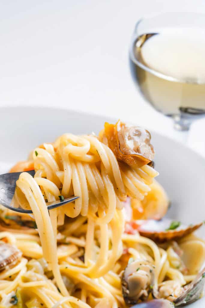 a forkful of spaghetti with a clam and a plate of spaghetti with clam sauce in the background.