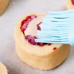 A close-up of a raw scone with raspberries on parchment paper being brushed with milk using a blue silicone pastry brush.