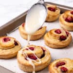 A spoon drizzling white glaze over freshly baked raspberry scones on a parchment-lined baking tray.