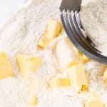 Cubed butter being blended into flour with a pastry cutter in a clear glass bowl.