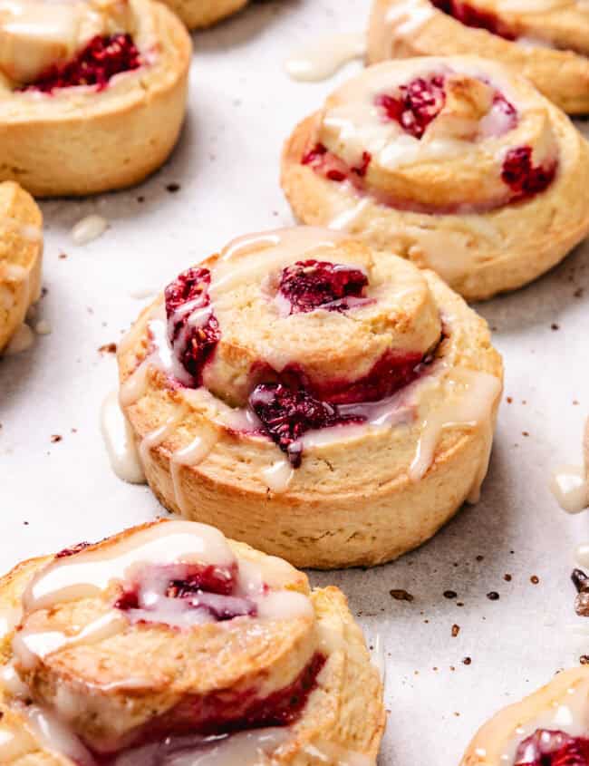 Several Raspberry swirled Scones with white glaze on top are neatly arranged on parchment paper. Raspberries and a spoon with glaze are scattered around them.