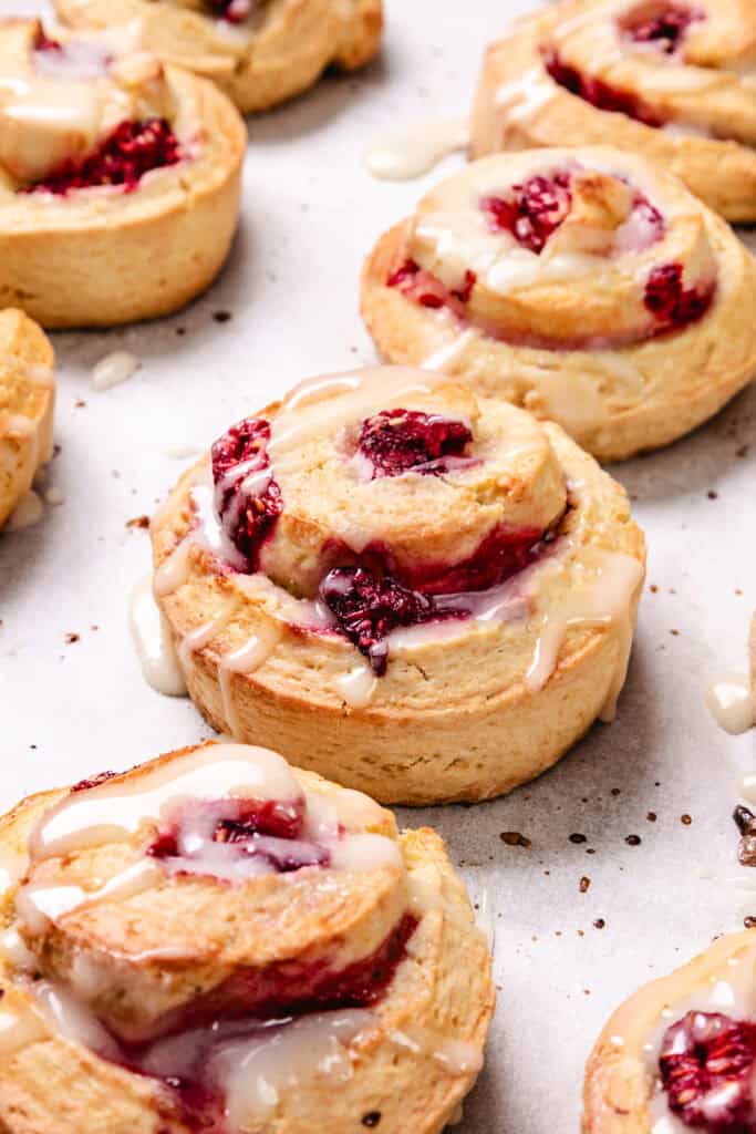 Several Raspberry swirled Scones with white glaze on top are neatly arranged on parchment paper. Raspberries and a spoon with glaze are scattered around them.