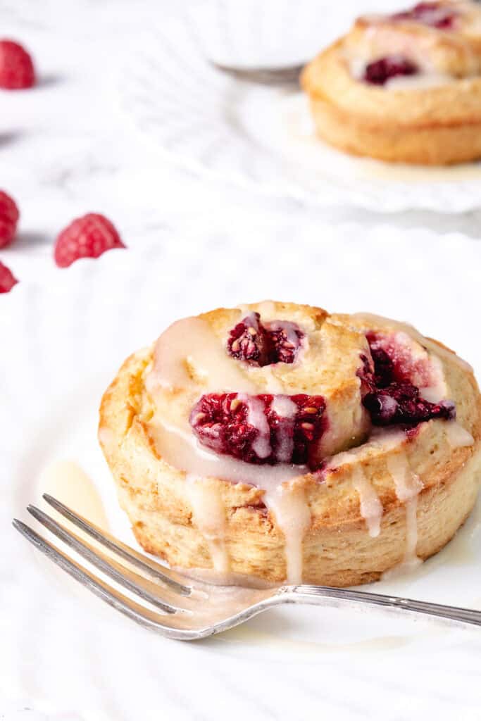 A Raspberry Scone swirled with raspberries and a drizzle of icing on a white surface, with a fork and extra raspberries nearby.