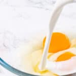 Cream being poured into a clear glass bowl with a whole egg, set on a marble countertop.