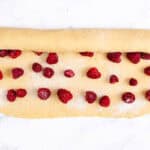 Scone dough on a marble surface, rolled halfway to enclose scattered fresh raspberries for filling.