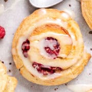 A top-down view of golden-brown scones with raspberries and icing, surrounded by individual raspberries on parchment paper.