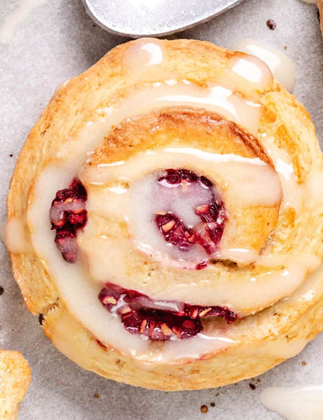 A top-down view of golden-brown scones with raspberries and icing, surrounded by individual raspberries on parchment paper.