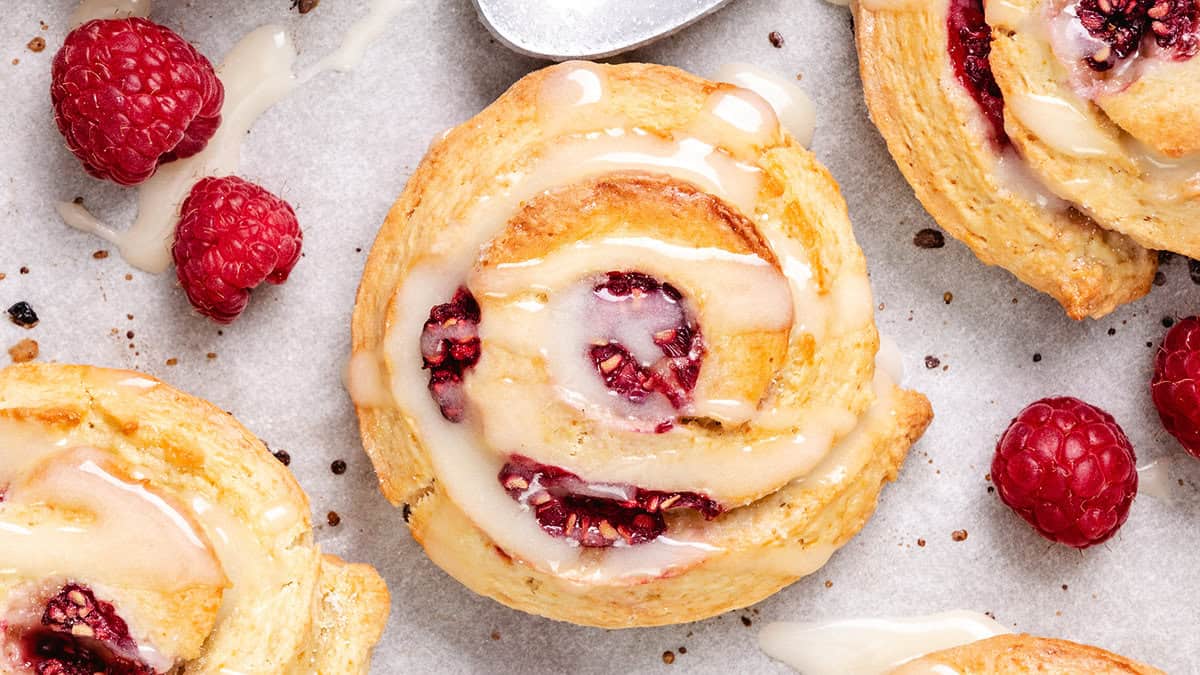 A top-down view of golden-brown scones with raspberries and icing, surrounded by individual raspberries on parchment paper.