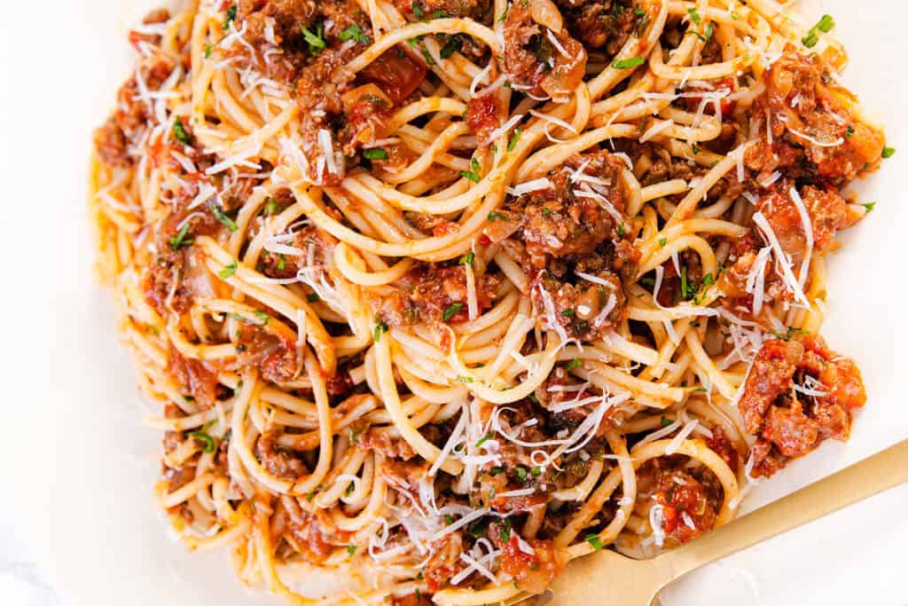 A close-up of a plate of quick & easy spaghetti Bolognese topped with minced meat sauce and garnished with freshly grated Parmesan cheese. A gold-colored fork is placed alongside the pasta.