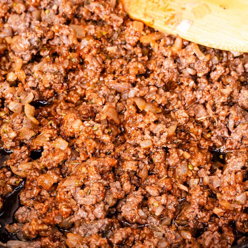 Close-up of seasoned ground meat with diced onions, perfect for a Quick & Easy Spaghetti Bolognese, being cooked in a pan. A wooden spatula is partially visible, stirring the mixture, now slightly browned and textured with herbs and spices.