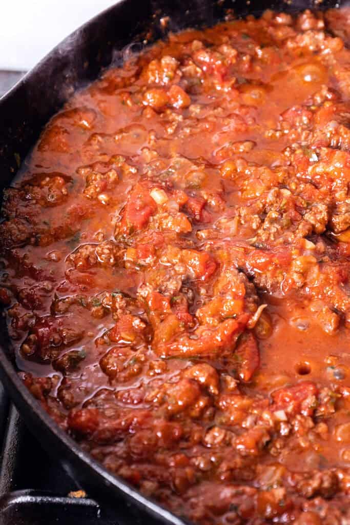 Close-up of a skillet filled with rich, chunky tomato meat sauce simmering on the stove, perfect for a quick & easy Spaghetti Bolognese. The sauce boasts visible pieces of ground meat, tomatoes, and herbs, creating a hearty and savory texture. Steam is rising from the bubbling mixture.