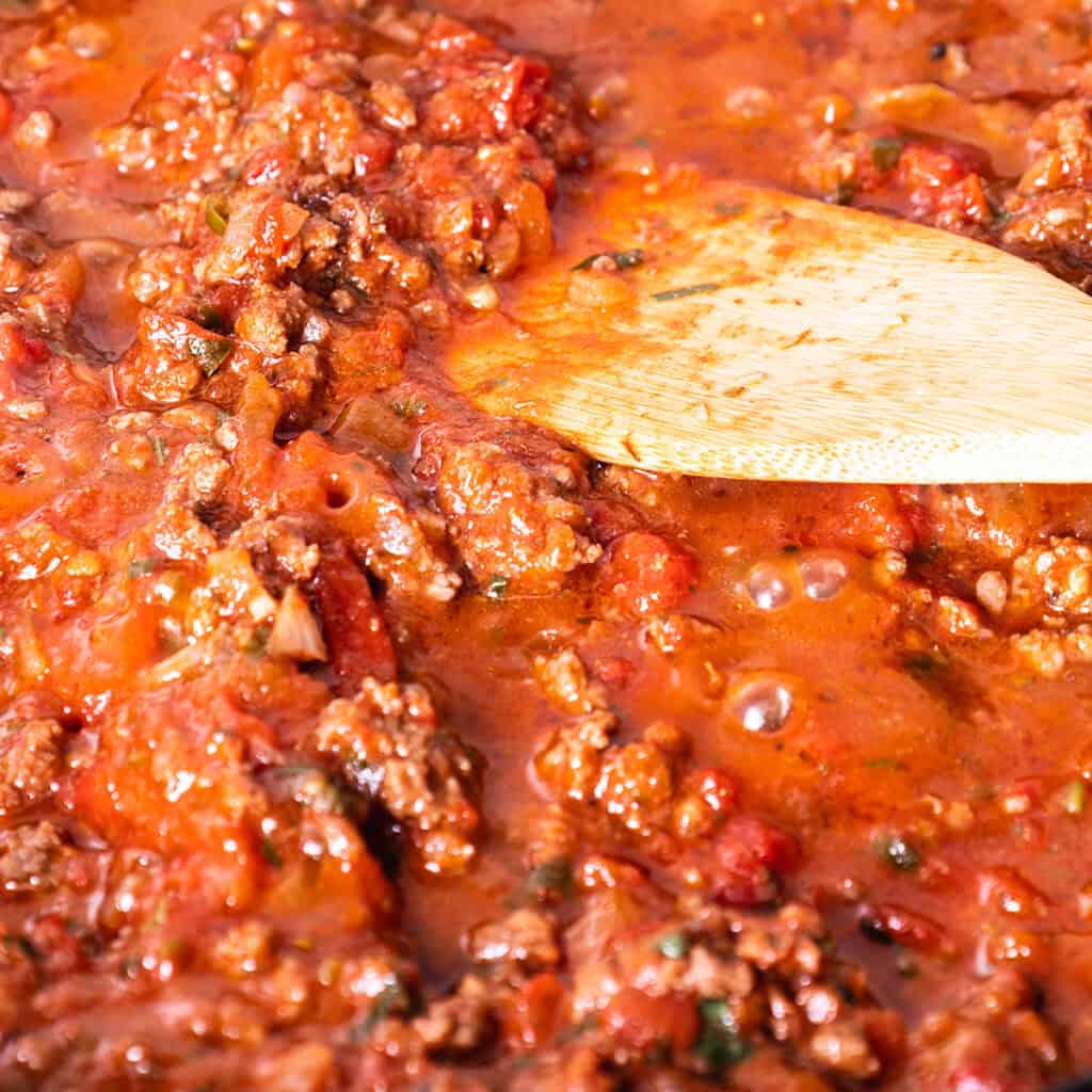 A wooden spoon stirs a rich, red tomato and ground beef sauce with visible chunks of tomato and herbs, simmering in a pan—a quick and easy Spaghetti Bolognese waiting to delight your taste buds.