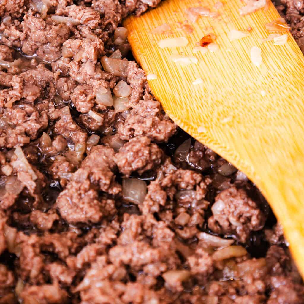 Close-up of ground beef being cooked with chopped onions in a pan, the beginning of a Quick & Easy Spaghetti Bolognese. A wooden spatula stirs the mixture, revealing browned textures and translucent onions. Steam rises, indicating the dish is hot and in progress.