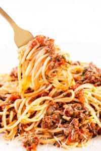 A fork lifts a swirl of quick & easy spaghetti bolognese from a plate. The pasta is richly coated in a tomato-based meat sauce, with visible herbs and grated cheese, creating a savory and comforting dish.