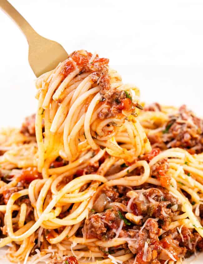 A fork lifts a swirl of quick & easy spaghetti bolognese from a plate. The pasta is richly coated in a tomato-based meat sauce, with visible herbs and grated cheese, creating a savory and comforting dish.