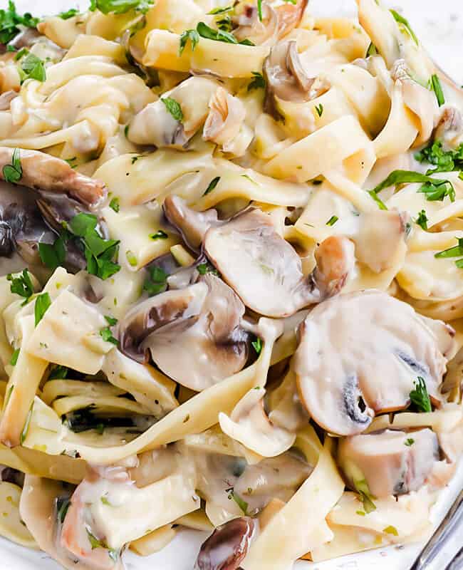 A close-up photo of a white plate overflowing with creamy tagliatelle pasta and sliced cremini mushrooms. A silver fork rests on the side of the plate.