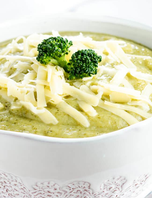 An image showing a white bowl filled with broccoli soup, garnished with shredded cheese, up close.