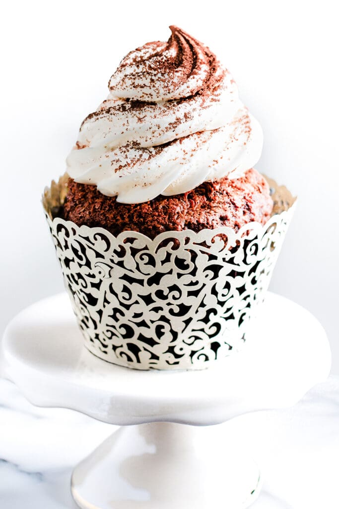 A close-up of a chocolate cupcake on cake stand. The cupcake is frosted with whipped cream and dusted with cocoa powder with a white background.