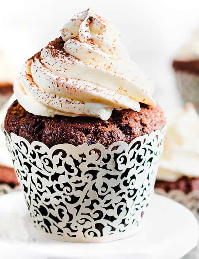 A close-up of two chocolate cupcakes on a white plate. The cupcakes are topped with whipped cream frosting and dusted with cocoa powder.