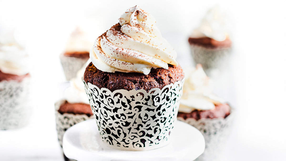 A close-up of two chocolate cupcakes on a white plate. The cupcakes are topped with whipped cream frosting and dusted with cocoa powder.