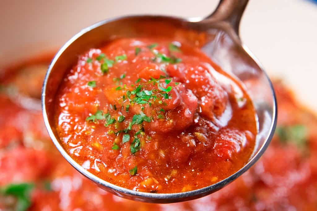 A close-up of a ladle filled with vibrant tomato sauce, garnished with fresh parsley. The chunky texture highlights pieces of tomato. In the background, a blur of rich red sauce simmers in the pot.