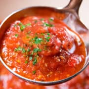 A close-up of a ladle filled with vibrant tomato sauce, garnished with fresh parsley. The chunky texture highlights pieces of tomato. In the background, a blur of rich red sauce simmers in the pot.