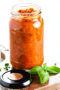 A jar of Tomato Sauce with basil and chopped parsley scattered around it