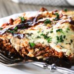 A slice of Moussaka on a white plate and a fork next to it