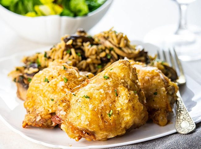 Crispy Oven Baked Chicken Thighs on a white plate with rice and salad in the background