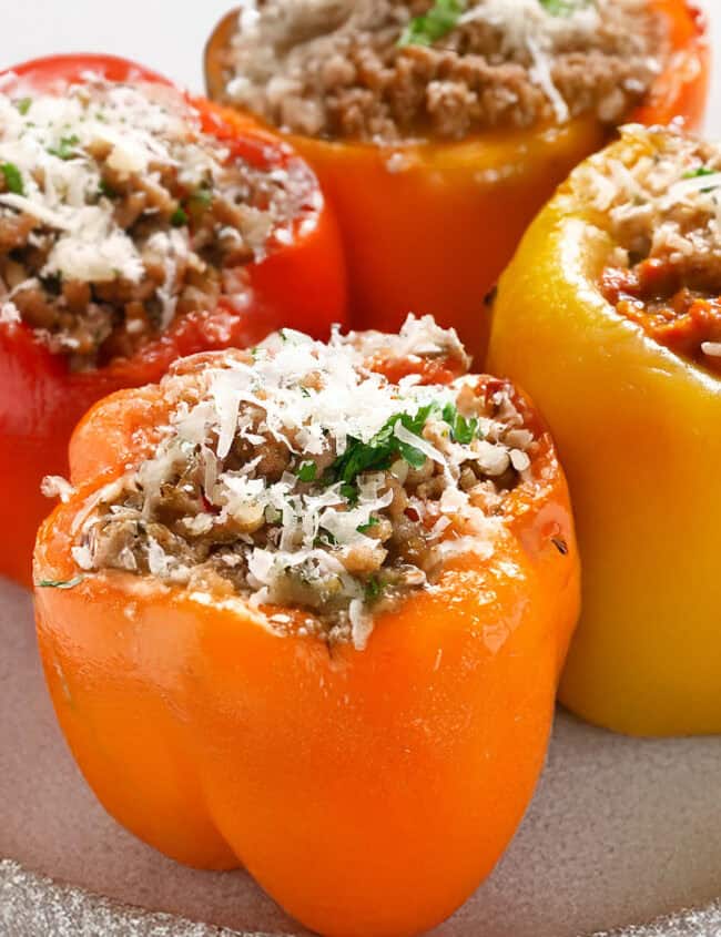 Four Italian Stuffed Peppers filled with seasoned ground meat and topped with melted cheese. The peppers are red, orange, and yellow, garnished with chopped herbs, and are placed on a white plate.