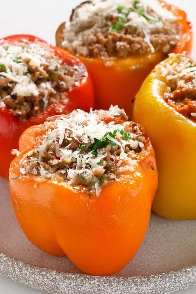 Four Italian Stuffed Peppers filled with seasoned ground meat and topped with melted cheese. The peppers are red, orange, and yellow, garnished with chopped herbs, and are placed on a white plate.