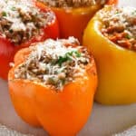 Four Italian stuffed bell peppers in red, orange, and yellow sit on a round white plate. They are filled with a savory mixture of rice, minced meat, herbs, and topped with grated cheese. The vibrant colors create a visually appealing dish that's as delightful to taste as it is to behold.