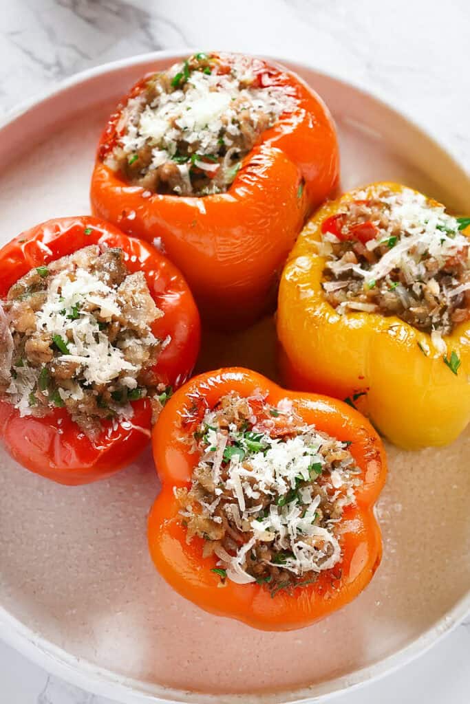 Four Italian stuffed bell peppers filled with rice, ground meat, and savory herbs are topped with grated cheese. These vibrant red, yellow, and orange delights are elegantly arranged on a round, white plate.