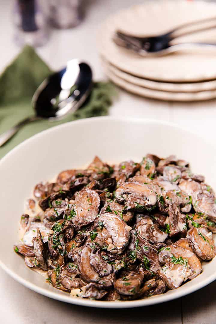 a bowl piled high with creamy garlic mushrooms sprinckled with parsley with plates, forks and a serving spoon in the background 