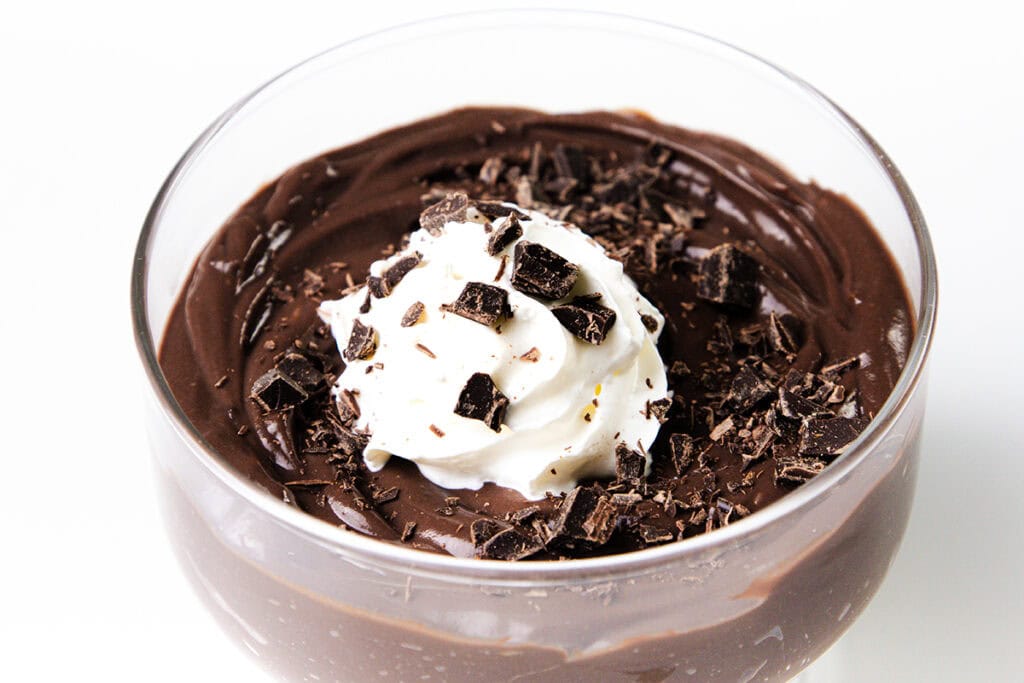 A glass cup brimming with luscious chocolate pudding is topped with a swirl of whipped cream and garnished with dark chocolate shavings, set against a plain white background.