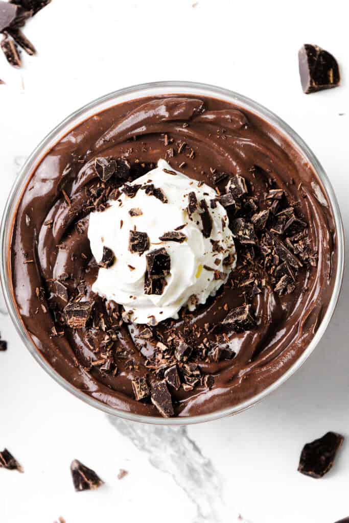 A bowl of luscious chocolate pudding topped with a swirl of whipped cream and sprinkled with dark chocolate shavings rests on a white marble surface, with extra chocolate pieces scattered around.