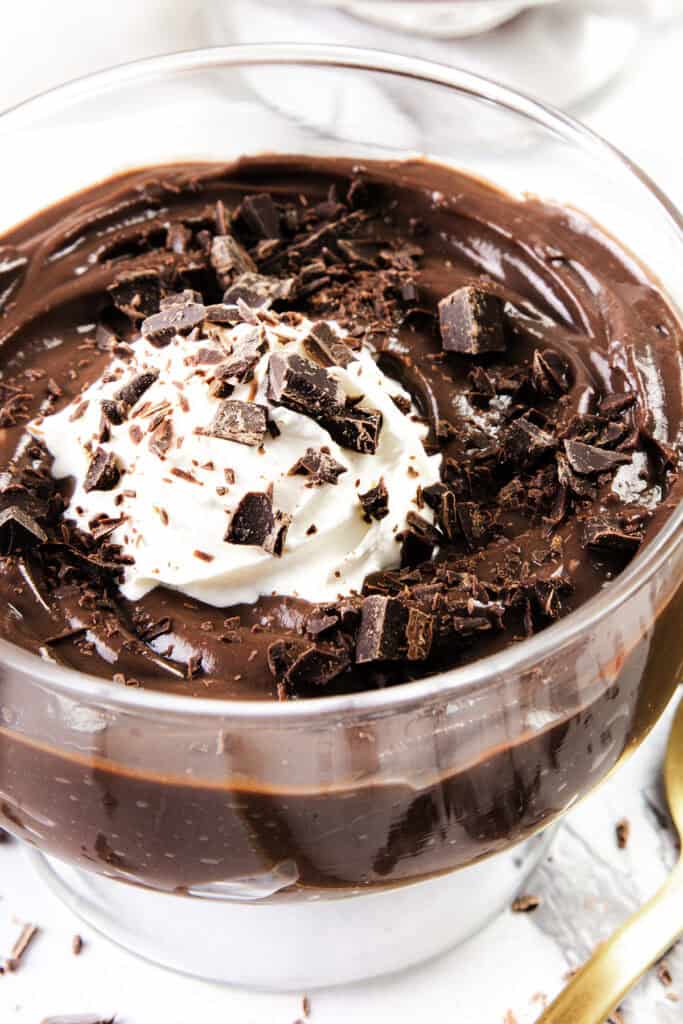 A glass bowl brimming with decadent chocolate pudding, crowned by a dollop of whipped cream and drizzled in delicate dark chocolate shavings, rests elegantly against a light backdrop.