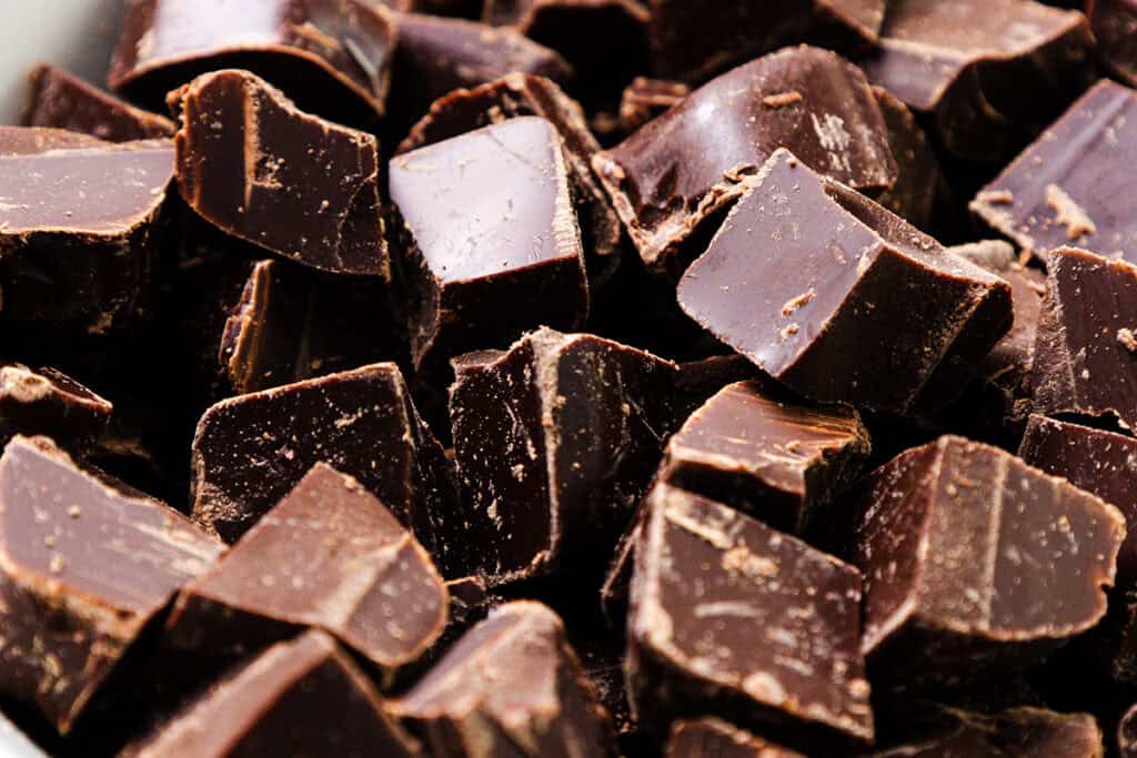 Close-up of a pile of dark chocolate chunks, reminiscent of indulgent chocolate pudding. The pieces have a glossy surface with some rough edges and varying sizes, capturing the rich texture of the chocolate.