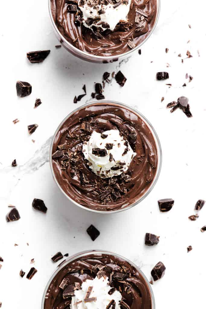 Three glass cups filled with rich chocolate mousse and velvety chocolate pudding are topped with whipped cream and sprinkled with chocolate shavings, surrounded by scattered pieces of chopped chocolate on a white marble surface.