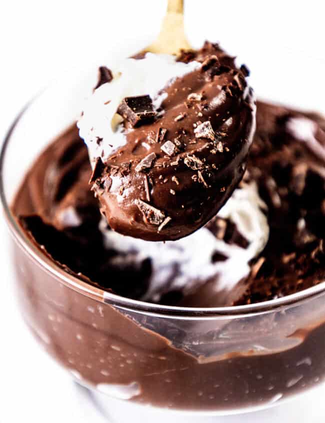 Close-up of a glass bowl filled with rich, creamy chocolate mousse and topped with whipped cream and chocolate shavings. A spoon lifts a generous scoop, revealing the smooth texture of this decadent chocolate pudding delight.