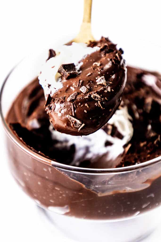 Close-up of a glass bowl filled with rich, creamy chocolate mousse and topped with whipped cream and chocolate shavings. A spoon lifts a generous scoop, revealing the smooth texture of this decadent chocolate pudding delight.