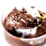 A close-up of a spoonful of decadent chocolate pudding, adorned with delicate chocolate shavings and a swirl of whipped cream. The spoon hovers tantalizingly above a glass brimming with more luscious chocolate pudding, set against a bright, blurred background.