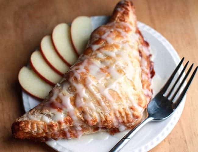 A crispy. golden brown apple turnover on a plate drizzled with icing. A fork and some sliced apples are beside it on the plate