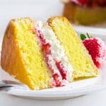 A slice of Lemon Sponge Cake filled with whipped cream and strawberries on a plate with the cake behind it