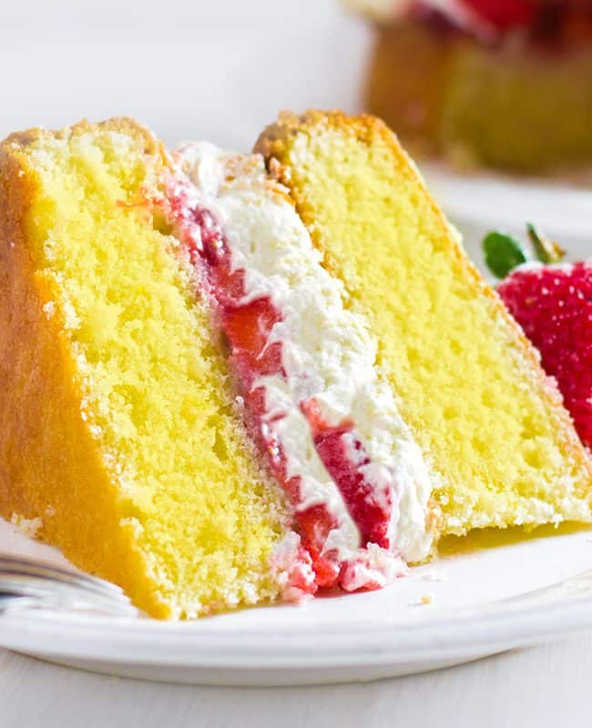 A slice of Lemon Sponge Cake filled with whipped cream and strawberries on a plate with the cake behind it