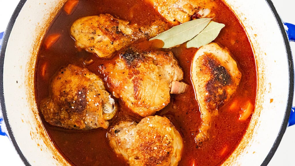 A pot of chicken stew with browned pieces, visible carrots, and a rich red broth reminiscent of Coq Au Vin. A bay leaf rests on top, adding a touch of garnish to the savory dish in the white pot with a blue rim.