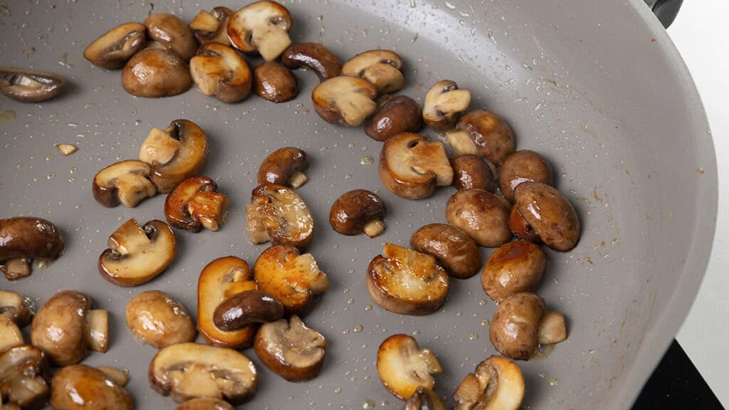 Sliced brown mushrooms sautéing in a non-stick pan, glistening with oil, add a rustic Coq Au Vin touch.